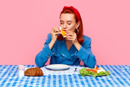 Photo for Food pop art photography. Young redhead woman sitting at table with checkered tablecloth and eating breakfast with fried eggs against pink studio background. Complementary colors. Copy space for ad - Royalty Free Image