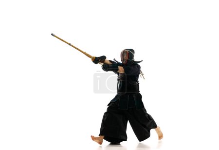 Téléchargez les photos : Homme, athlète professionnel de kendo en mouvement, en uniforme d'entraînement avec épée de bambou, shinai sur fond de studio blanc. Concept d'arts martiaux, sport, culture japonaise, action et mouvement - en image libre de droit