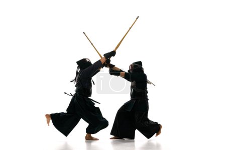Téléchargez les photos : Image dynamique de deux hommes, athlètes professionnels de kendo en uniforme avec casque d'entraînement avec épée en bambou shinai sur fond de studio blanc. Concept d'arts martiaux, sport, culture japonaise, action - en image libre de droit