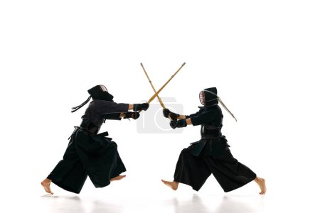 Téléchargez les photos : Image dynamique de deux hommes, des athlètes professionnels de kendo s'entraînant avec l'épée de bambou shinai sur fond de studio blanc. Concept d'arts martiaux, sport, culture japonaise, action et mouvement - en image libre de droit