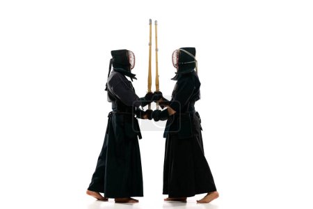 Full-length image of two men, professional kendo fighter, athletes in black uniform training against white studio background. Concept of martial arts, sport, Japanese culture, action and motion