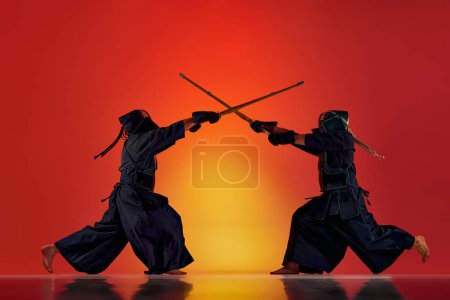 Téléchargez les photos : Image dynamique de deux hommes, des athlètes professionnels de kendo s'entraînant avec l'épée de bambou shinai contre fond de studio rouge dégradé au néon. Concept d'arts martiaux, sport, culture japonaise, mouvement - en image libre de droit