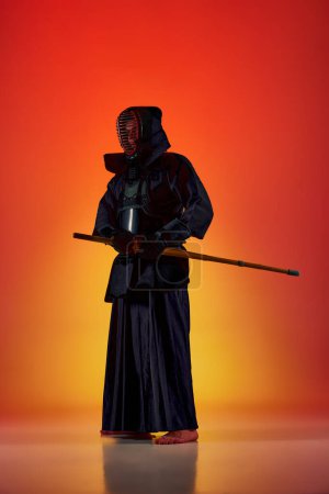 Téléchargez les photos : Portrait complet de l'homme, athlète professionnel de kendo en uniforme posant avec l'épée de shinai contre fond de studio rouge dégradé en lumière au néon. Arts martiaux, sport, culture japonaise, concept de mouvement - en image libre de droit
