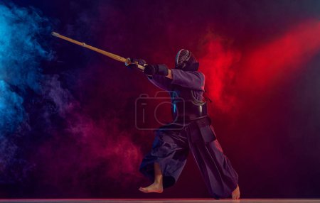 Foto de Hombre, atleta kendo profesional, deportista de combate posando con espada shinai contra el degradado fondo oscuro en luz de neón con humo. Concepto de artes marciales, deporte, cultura japonesa, acción y - Imagen libre de derechos