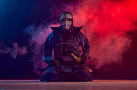 Téléchargez les photos : Homme, athlète professionnel de kendo, sportif de combat posant avec l'épée shinai contre fond sombre dégradé dans la lumière au néon avec de la fumée. Concept d'arts martiaux, sport, culture japonaise, action et - en image libre de droit