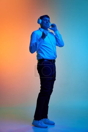 Téléchargez les photos : Portrait complet d'un homme d'âge moyen positif écoutant de la musique dans un casque et dansant sur fond bleu orange dégradé sous la lumière du néon. Concept d'émotions, style de vie, expression faciale. Publicité - en image libre de droit