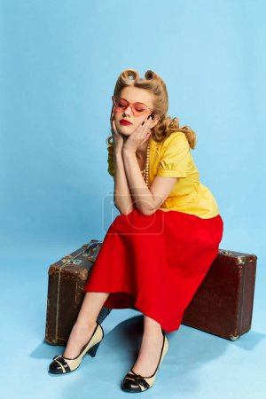 Téléchargez les photos : Portrait de belle jeune fille en chemisier jaune et jupe rouge assise sur des valises vintage sur fond de studio bleu. Concept de mode rétro, beauté, voyage, années 50, 60. Style pin-up - en image libre de droit