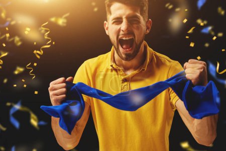 Photo for Positivity. Young man, sport fan emotionally cheering up favourite Ukrainian football team over dark background with confetti. Concept of sport, leisure time, emotions, hobby and entertainment - Royalty Free Image