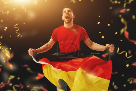 Téléchargez les photos : Jeune homme, fan de football encourageant émotionnellement l'équipe allemande préférée sur fond sombre avec des confettis. Soutien. Concept de sport, temps libre, émotions, passe-temps et divertissement - en image libre de droit