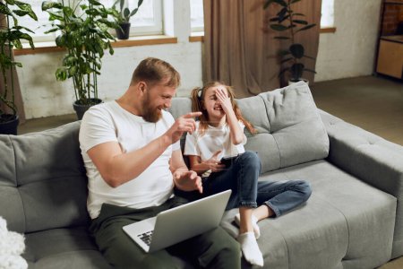 Photo for Happy smiling mature man, father sitting on couch with laptop, working at home remotely and playing with his little daughter. Concept of fatherhood, childhood, family, freelance job, home office - Royalty Free Image