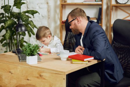 Photo for Mature man, father sitting at table, working remotely at home and doing homework with his little son. Attentive parent. Concept of fatherhood, childhood, family, freelance job, home office - Royalty Free Image