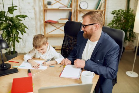 Photo for Mature man, father sitting at table, working remotely at home and doing homework with his little son. Attentive parent. Concept of fatherhood, childhood, family, freelance job, home office - Royalty Free Image