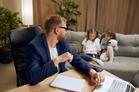 Photo for Mature man, father sitting at table and working on laptop remotely at home and looking after his little children playing on sofa. Concept of fatherhood, childhood, family, freelance job, home office - Royalty Free Image