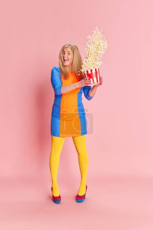 Téléchargez les photos : L'heure du film. Portrait complet de femme en vêtements colorés posant émotionnellement avec du pop-corn sur fond de studio rose. Concept d'émotions, expression faciale, mode de vie et mode - en image libre de droit
