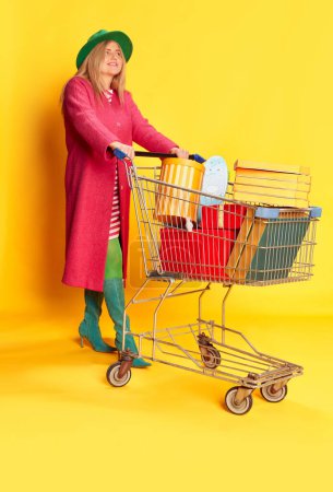 Photo for Portrait of woman in colorful clothes, pink coat and green hat posing with shopping trolley against yellow studio background. Sales. Concept of emotions, facial expression, lifestyle and fashion - Royalty Free Image