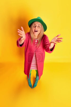 Photo for Top view. Portrait of emotive excited woman in colorful clothes, green hat and pink coat posing against yellow studio background. Concept of emotions, facial expression, lifestyle and fashion - Royalty Free Image