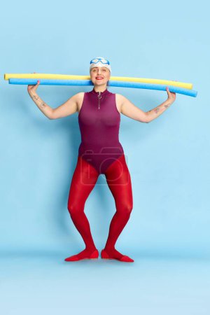 Photo for Full-length image of woman in swimming suit and red tights posing with swimming equipment against blue studio background. Concept of emotions, facial expression, lifestyle and fashion - Royalty Free Image
