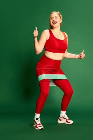 Photo for Full-length portrait of woman in red sportive clothes posing, training with fitness elastic bands against green studio background. Concept of emotions, facial expression, sport and weight loss - Royalty Free Image