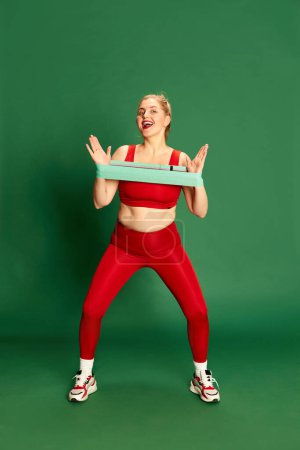 Téléchargez les photos : Portrait complet de femme en vêtements de sport rouge posant, entraînement avec des bandes élastiques de fitness sur fond de studio vert. Concept d'émotions, expression faciale, sport et perte de poids - en image libre de droit