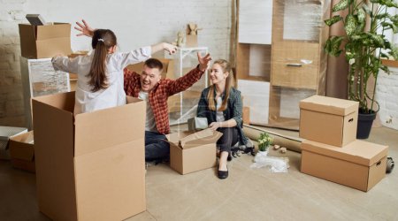 Photo for Young happy family, man, woman and kid moving into new flat, apartment with many cardboard boxes. Father playing with little daughter. Concept of moving houses, real estate, family, new life - Royalty Free Image