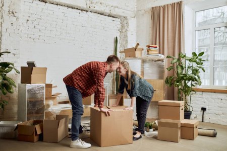 Photo for Happy young family, man and woman moving into new house, apartment. Opening many cardboard boxes. Furnishing room on daytime. Concept of moving houses, real estate, family, new life - Royalty Free Image