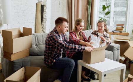 Photo for Putting things on place. Young happy family, man, woman and kid moving into new flat, apartment with many cardboard boxes. Concept of moving houses, real estate, family, new life - Royalty Free Image