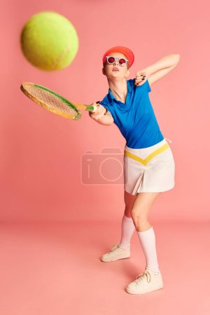 Téléchargez les photos : Frapper la balle. Portrait de belle jeune fille en vêtements de sport posant avec raquette de tennis sur fond rose. Concept de jeunesse, sport, mode, mode de vie, émotions, expression faciale. Publicité - en image libre de droit