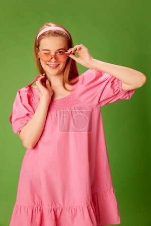 Téléchargez les photos : Jolie tenue. Portrait de belle jeune fille blonde en robe rose mignonne et lunettes de soleil sur fond de studio vert. Concept de jeunesse, beauté, mode, mode de vie, émotions, expression faciale. Publicité - en image libre de droit