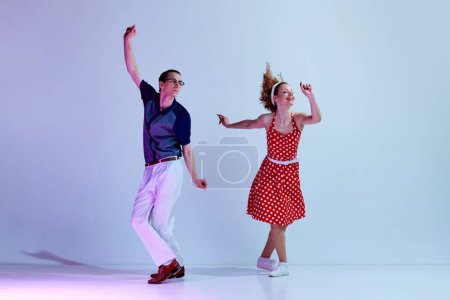 Téléchargez les photos : Jeune couple positif, homme et femme en costumes colorés dansant des danses de style rétro sur fond de studio pourpre bleu dégradé. Concept d'art, style rétro, passe-temps, fête, plaisir, années 60, 70 culture - en image libre de droit