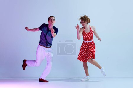 Foto de Hermosa pareja joven, hombre y mujer con trajes elegantes bailando bailes retro contra el fondo del estudio azul púrpura degradado. Arte, estilo retro, hobby, fiesta, diversión, movimientos, 60s, 70s culture concept - Imagen libre de derechos