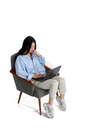 Téléchargez les photos : Vue isométrique supérieure. Jeune fille en chemise assise sur une chaise et travaillant, étudiant dans un ordinateur portable isolé sur fond de studio blanc. Concept d'entreprise, d'emploi, d'éducation. style de vie. Espace de copie pour la publicité - en image libre de droit