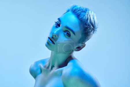 Photo for Beautiful young girl with short blonde hair and piercing posing with bare shoulders against blue studio background in neon light. Cyberpunk style. Concept of technology, modern fashion, digital art - Royalty Free Image
