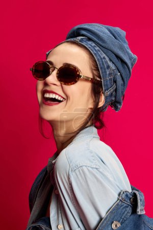 Foto de Felicidad, positiva. Retrato de una joven hermosa mujer sonriendo, posando con un elegante atuendo de vaqueros sobre fondo rojo del estudio. Concepto de estilo extraordinario, belleza, moda, juventud, emociones. Anuncio - Imagen libre de derechos