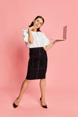 Foto de Retrato de larga duración de la joven mujer de negocios hermosa en ropa formal posando con el ordenador portátil sobre fondo de estudio rosa. Concepto de negocio, educación, ocupación, emociones, moda. Anuncio - Imagen libre de derechos
