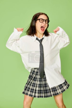 Téléchargez les photos : Jeune femme émotionnelle et énergique en jupe à carreaux et chemise blanche posant, écoutant de la musique dans des écouteurs sur fond de studio vert. Concept de jeunesse, mode, émotions, expression faciale - en image libre de droit