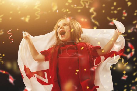 Téléchargez les photos : Petite fille, heureuse enfant avec drapeau d'Angleterre regardant émotionnellement le match de football au stade avec confettis. Gagner un match réussi. Concept de sport, coupe, monde, équipe, événement, compétition - en image libre de droit