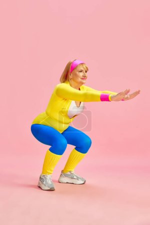 Téléchargez les photos : Sujet âgé belle femme dans la formation colorée de vêtements de sport, faire des squats, posant sur fond de studio rose. Rester en forme. Concept de vie sportive, retraite, soins de santé, bien-être. Publicité - en image libre de droit