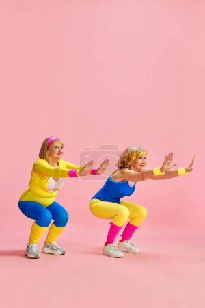 Téléchargez les photos : Deux sportives, senior, vieille femme dans la formation colorée de vêtements de sport, faire des squats, posant sur fond de studio rose. Concept de vie sportive, retraite, soins de santé, bien-être. Publicité - en image libre de droit