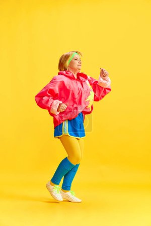 Photo for Full-length image of elderly sportive woman in colorful uniform training, running, posing against yellow studio background. Concept of sportive lifestyle, retirement, health care, wellness. Ad - Royalty Free Image