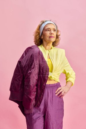 Foto de Hermosa mujer de mediana edad, deportiva en uniforme colorido posando sobre fondo de estudio rosa. Rutina fitness. Concepto de estilo de vida deportivo, jubilación, atención médica, bienestar. Anuncio - Imagen libre de derechos