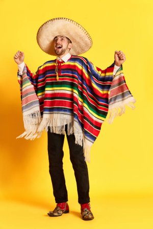 Téléchargez les photos : Portrait de jeune homme beau avec le visage heureux excité, dans des vêtements de fête colorés, poncho et sombrero posant sur fond jaune. Traditions mexicaines, célébration, festival, concept d'émotions - en image libre de droit