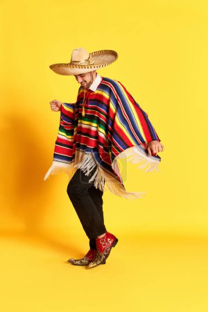 Téléchargez les photos : Portrait de jeune homme beau en vêtements de fête colorés, poncho et sombrero posant, dansant sur fond de studio jaune. Concept de traditions mexicaines, plaisir, célébration, festival, émotions - en image libre de droit