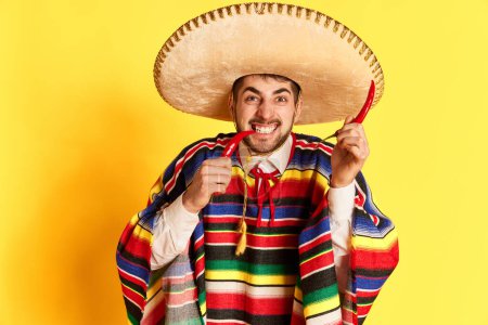 Téléchargez les photos : Jeune homme émotionnel en vêtements de fête colorés, poncho et sombrero posant avec du piment sur fond de studio jaune. Concept de traditions mexicaines, plaisir, célébration, festival, émotions - en image libre de droit