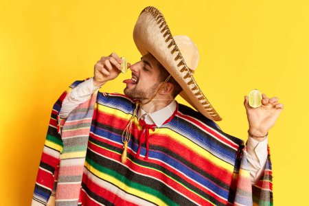 Téléchargez les photos : Jeune homme en poncho coloré et sombrero serrant de la chaux sur sa langue, posant sur fond de studio jaune. Concept de traditions mexicaines, plaisir, célébration, festival, émotions - en image libre de droit