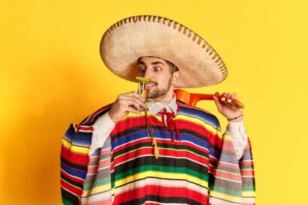 Téléchargez les photos : Jeune homme gai en poncho coloré et sombrero posant, reniflant la tequila à la chaux sur fond de studio jaune. Concept de traditions mexicaines, plaisir, célébration, festival, émotions - en image libre de droit