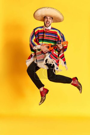 Photo for Young emotional man in colorful poncho and sombrero posing, playing guitar and jumping against yellow studio background. Concept of mexican traditions, fun, celebration, festival, emotions - Royalty Free Image