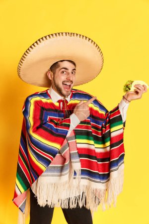 Photo for Portrait of young positive man in colorful clothes, poncho and sombrero posing, pointing at taco against yellow studio background. Concept of mexican traditions, fun, celebration, festival, emotions - Royalty Free Image