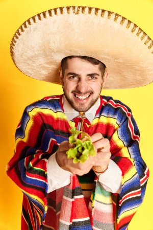 Photo for Portrait of young cheerful man in colorful festive clothes, poncho and sombrero posing wit taco against yellow studio background. Concept of mexican traditions, fun, celebration, festival, emotions - Royalty Free Image