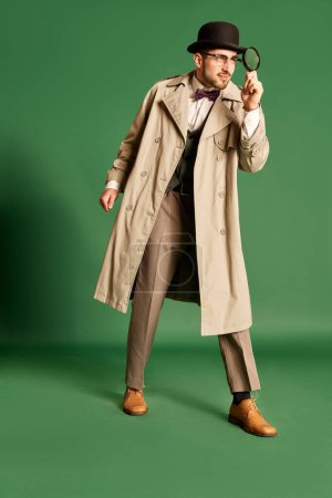 Photo for Full-length portrait of young man, detective in stylish trench coat and hat looking in magnifying glass against green studio background. Concept of emotions, profession, secrets, fun, fashion. Ad - Royalty Free Image