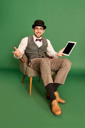 Téléchargez les photos : Portrait de jeune homme beau en vêtements classiques assis sur une chaise avec tablette, posant sur fond de studio vert. Concept d'émotions, profession de détective, secrets, amusement, mode. Publicité - en image libre de droit